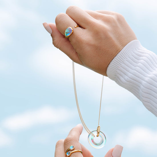 Hand holding a necklace with a clear pendant against a light blue sky.