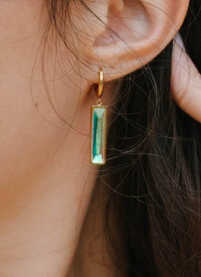 Woman wearing an Om Necklace with Bar Earrings, a white tank top and beige pants against a plain background