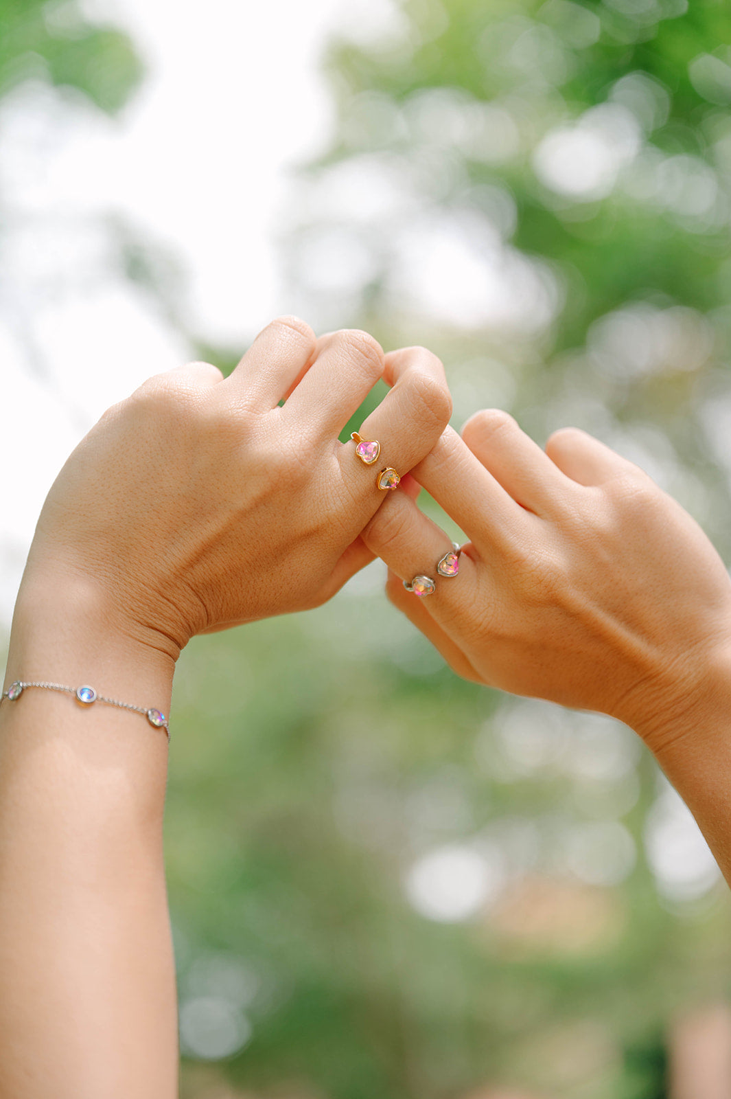 Two hands wearing heart rings  holding each other with blurred greenery in the background