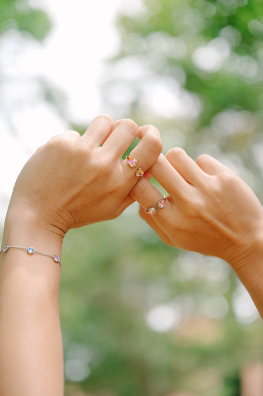Two hands wearing heart rings  holding each other with blurred greenery in the background